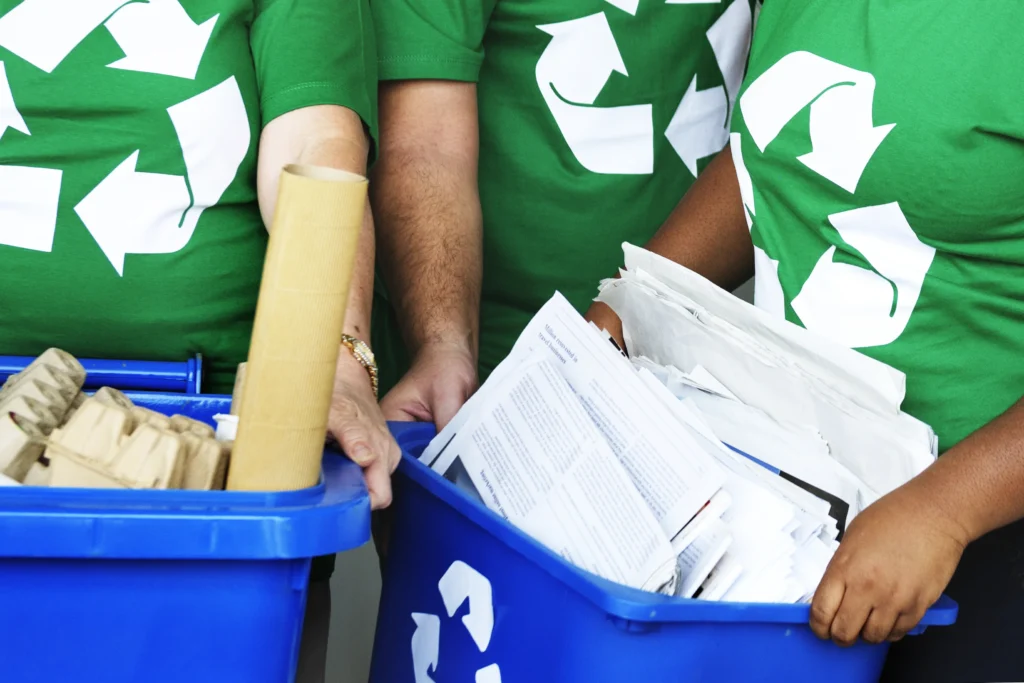 Des collaborateurs portant des t-shirt vert écologie avec le logo en gros du recyclage et portant des bacs de tri d'entreprise carton et papier pour sensibiliser les salariés à effectuer le tri des déchets sur leur lieu de travail.